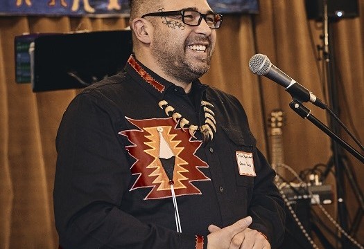 Aaron Dana, Passamaquoddy Tribal Representative in the Maine House of Representatives, speaks at the event. Aaron Dana, Passamaquoddy Tribal Representative in the Maine House of Representatives stands at a microphone smiling, hands clasped